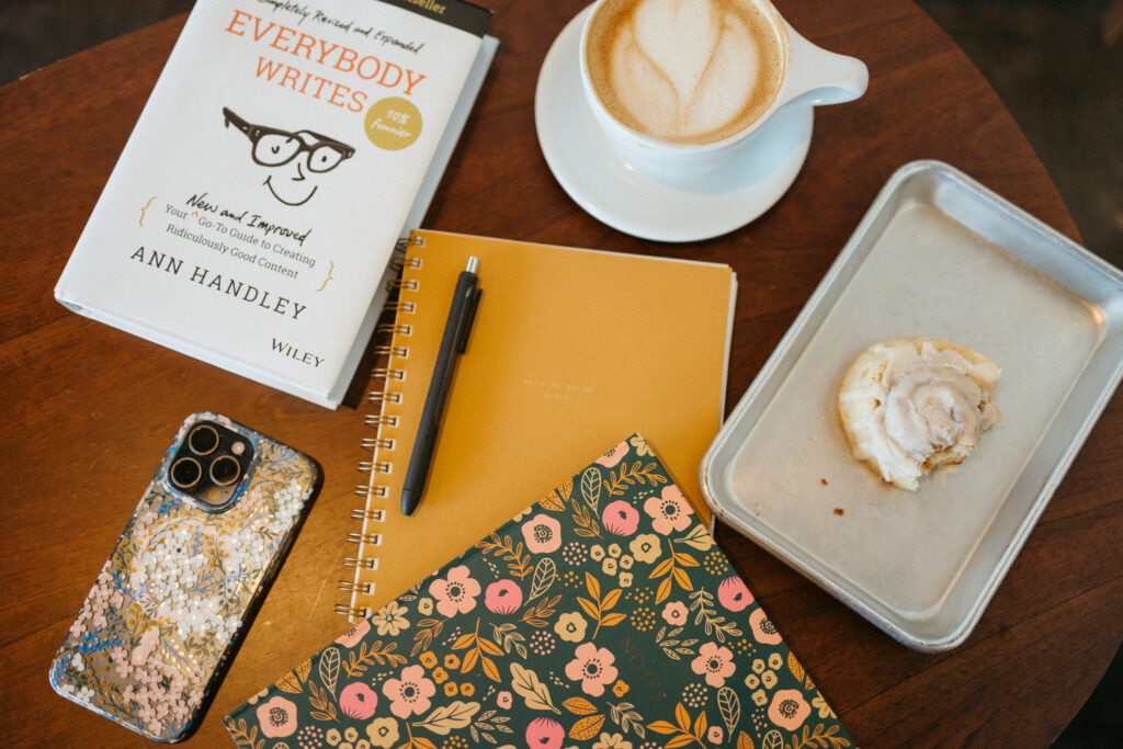 A coffee shop table with a book that teaches you how to write, a planner for blogging, and a notebook along with a cinnamon roll, coffee cup, and iphone for the use of blog content writer and SEO strategist Mckayla of The Comma Mama Co